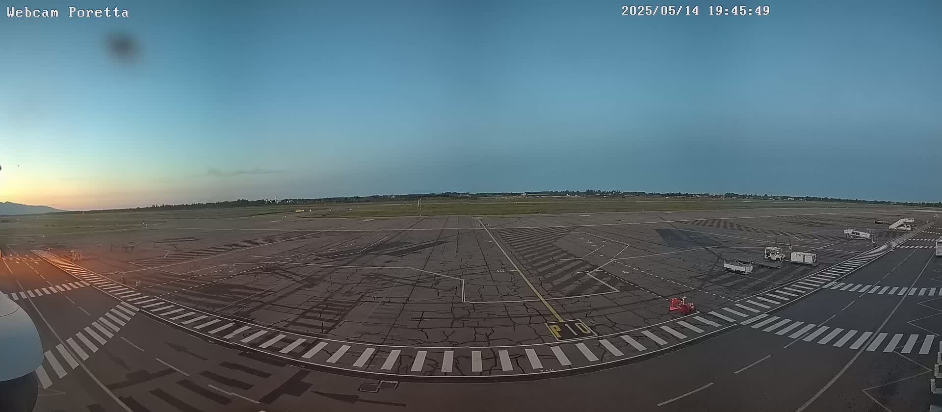 A mostly empty airport tarmac is viewed at dusk under a clear sky, with several service vehicles parked near the edge.