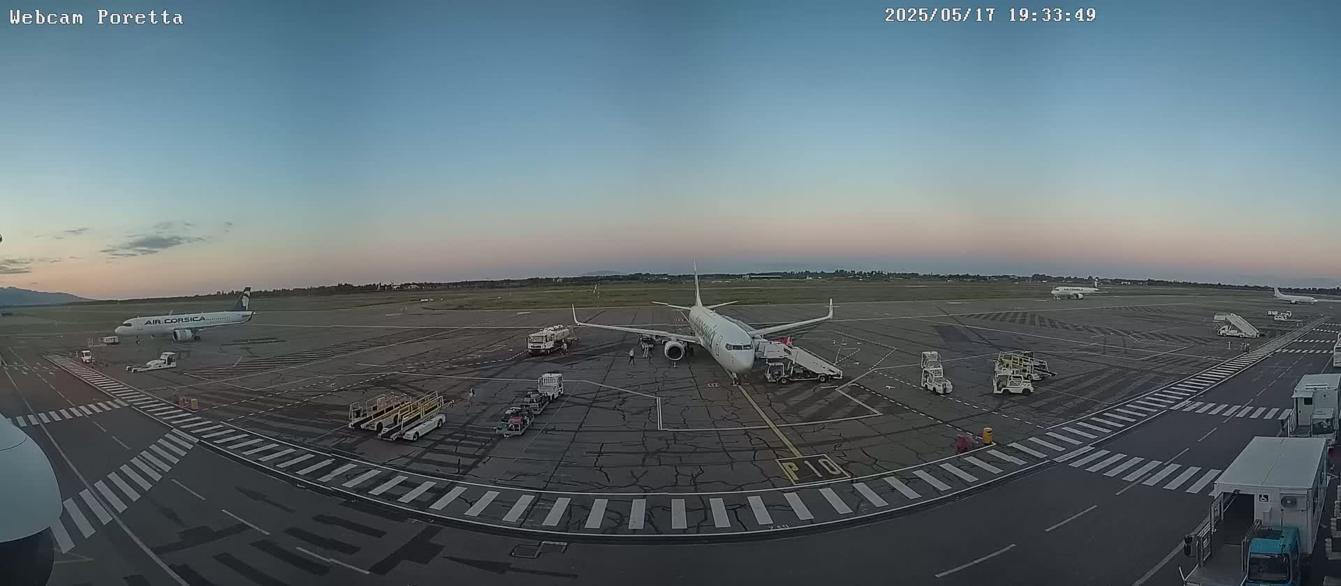 An airport tarmac at dusk shows several airplanes, ground support vehicles, and jet bridges under a clear, twilight sky.