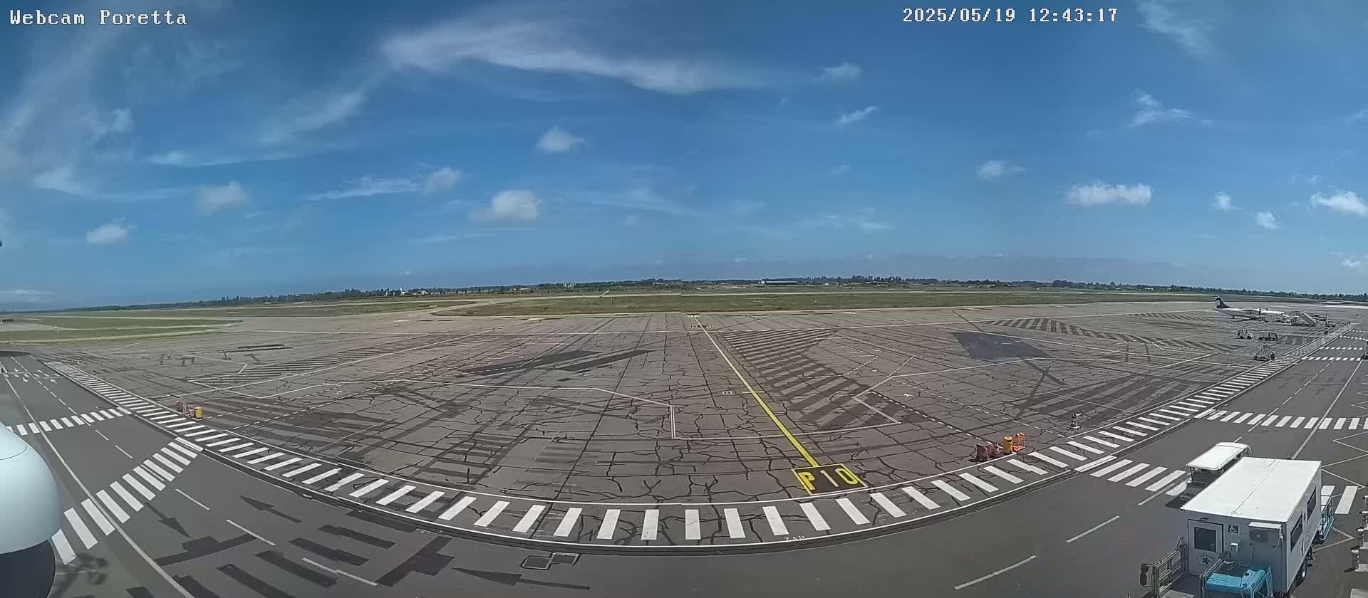 A mostly sunny day at an airport tarmac shows several planes parked and various ground support vehicles on the pavement.