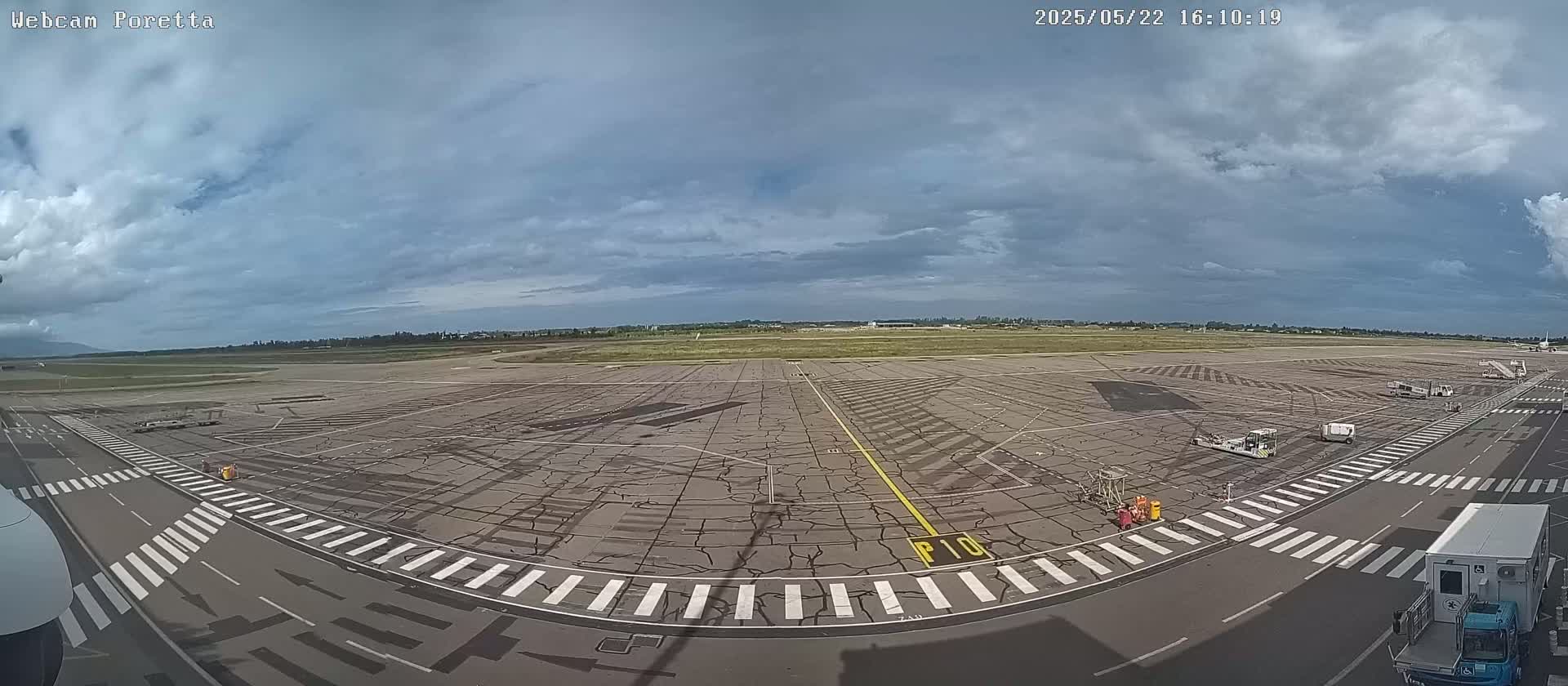 A wide view of an airport tarmac under a partly cloudy sky shows several ground service vehicles and a large expanse of cracked pavement with painted lines.