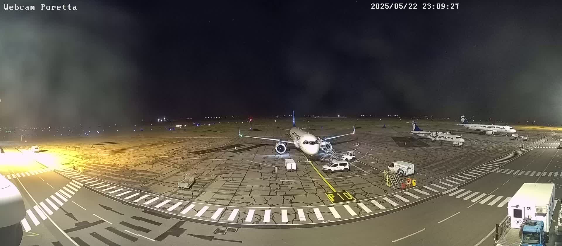 A nighttime airport tarmac shows several airplanes parked, ground support vehicles, and faintly visible lights in the distance under a mostly clear, dark sky.