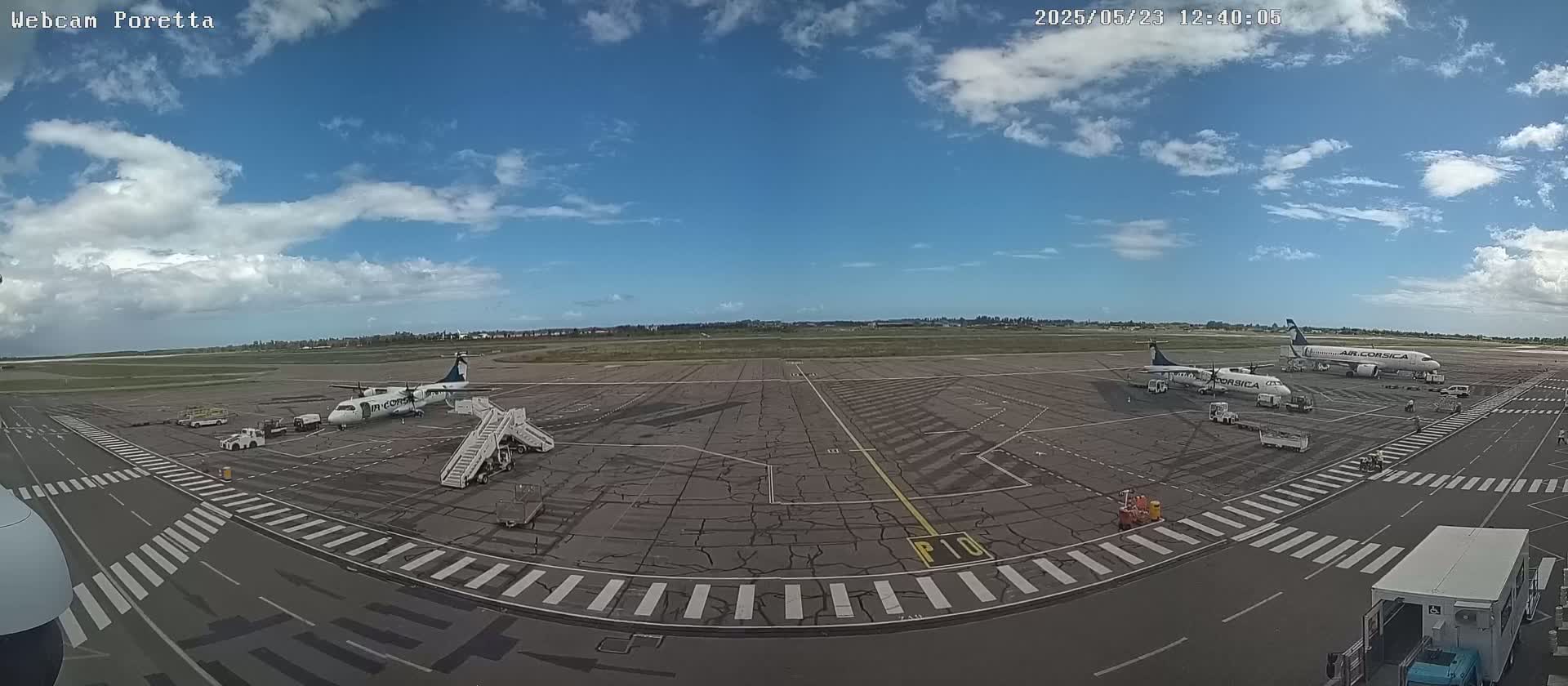 An airport tarmac under a partly cloudy sky shows several airplanes, ground support equipment, and marked roadways.