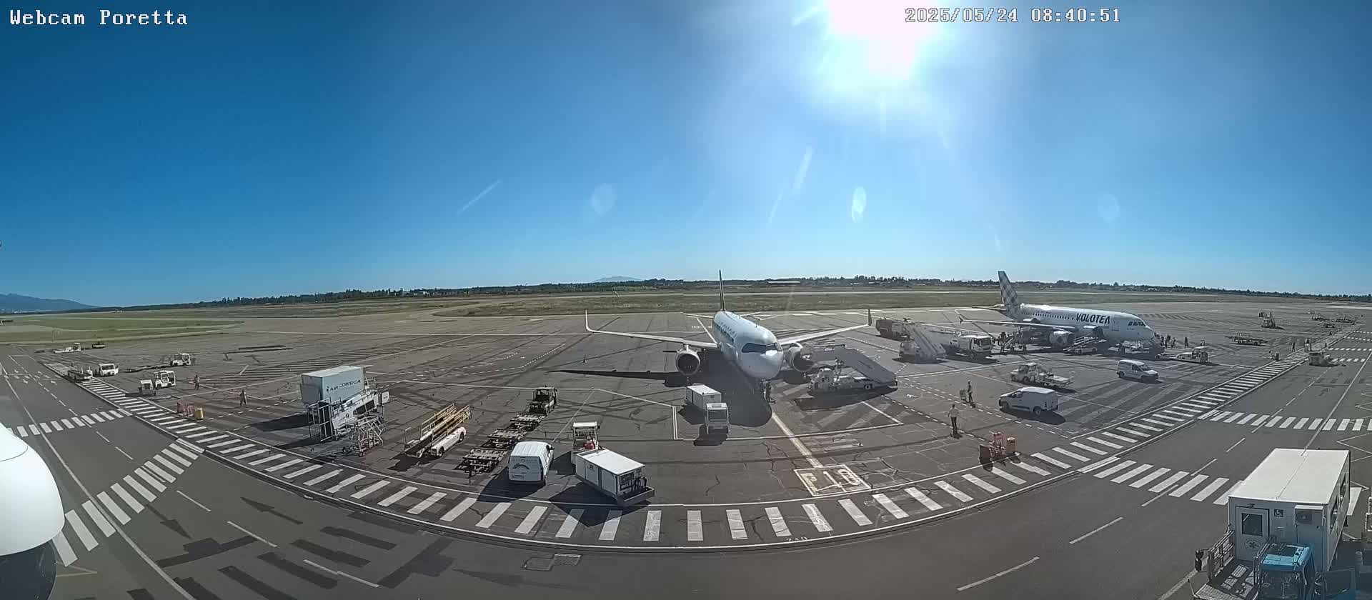 A sunny day at an airport shows two airplanes at the gate, surrounded by ground support vehicles and personnel.