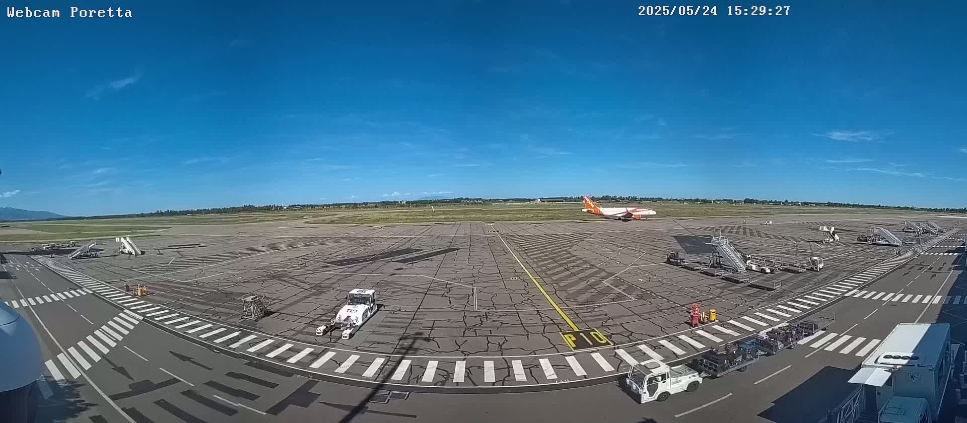 A mostly sunny airport tarmac with several airplanes and ground support equipment, including jet bridges and baggage carts.