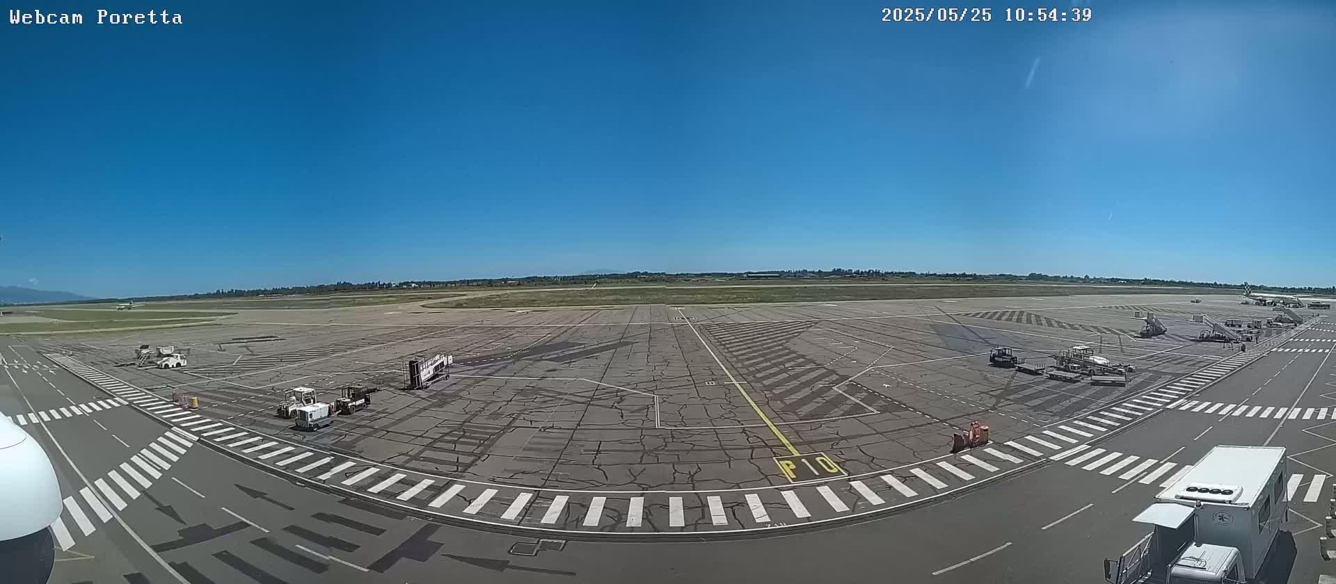 A mostly empty airport tarmac on a sunny day with several service vehicles and a few distant mountains visible on the horizon.