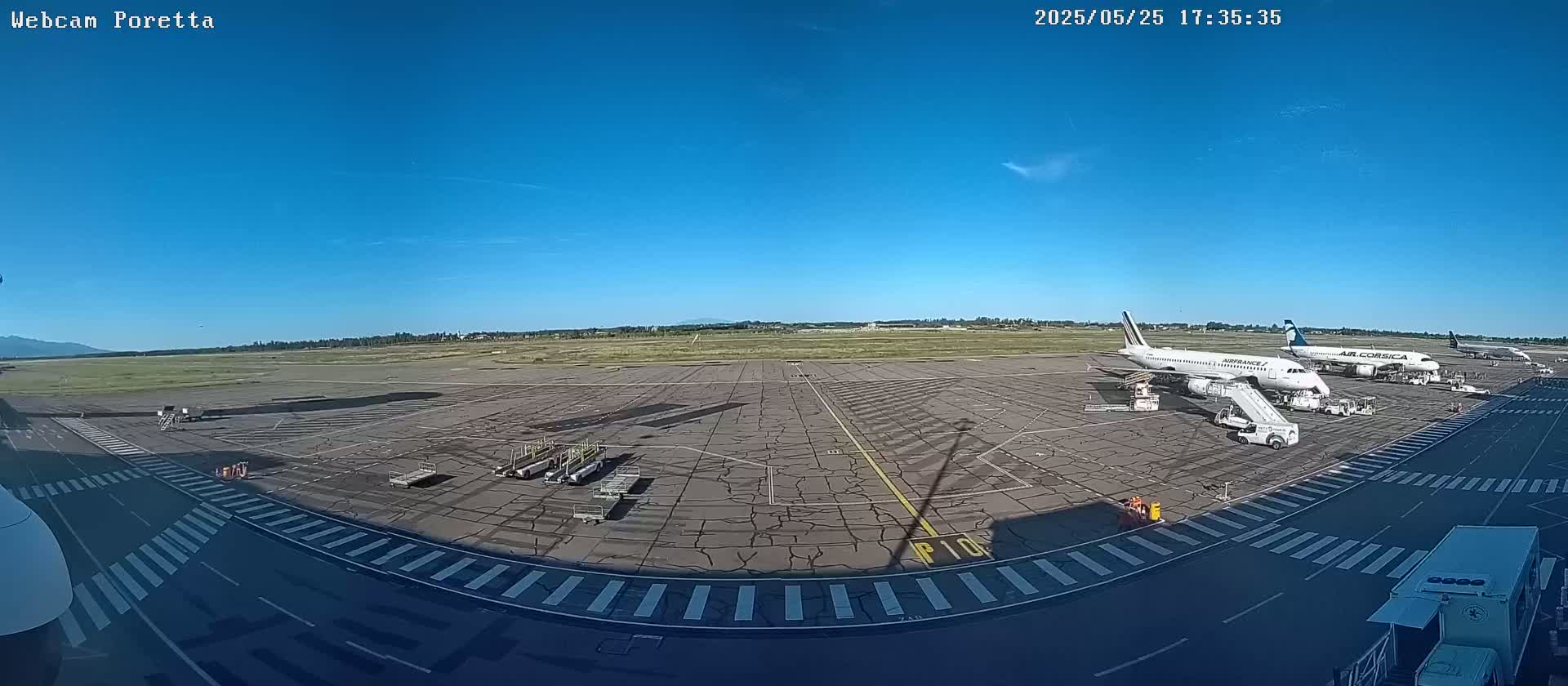 A wide shot of an airport tarmac on a sunny day shows several airplanes parked at gates with jet bridges attached, ground support equipment, and taxiways marked with stripes.