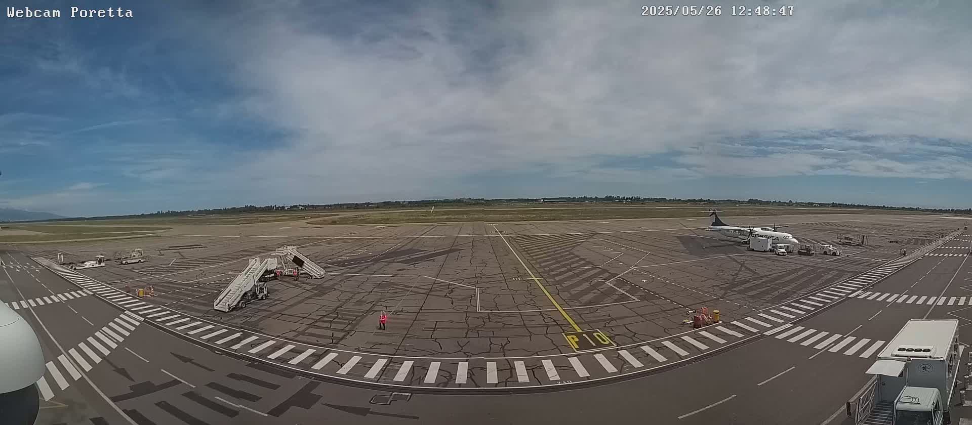 An airport tarmac under a partly cloudy sky shows a small passenger plane parked at a gate with jet bridges and ground support vehicles nearby.