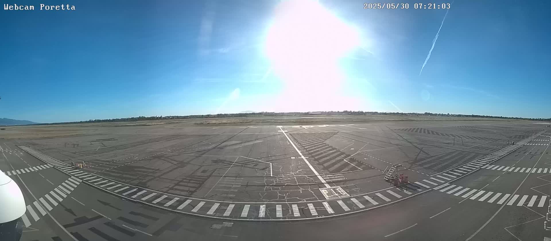 A sunny, mostly clear day reveals an empty airport tarmac with painted lines and crosswalks, viewed from a high angle.
