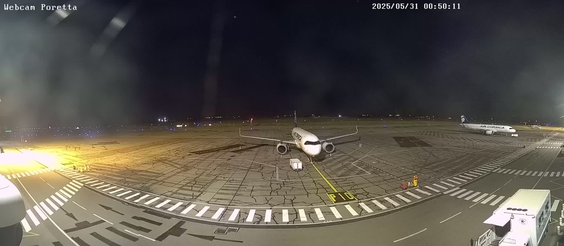 A nighttime view of an airport tarmac shows two airplanes parked, one in the foreground and another in the background, under a clear, dark sky.