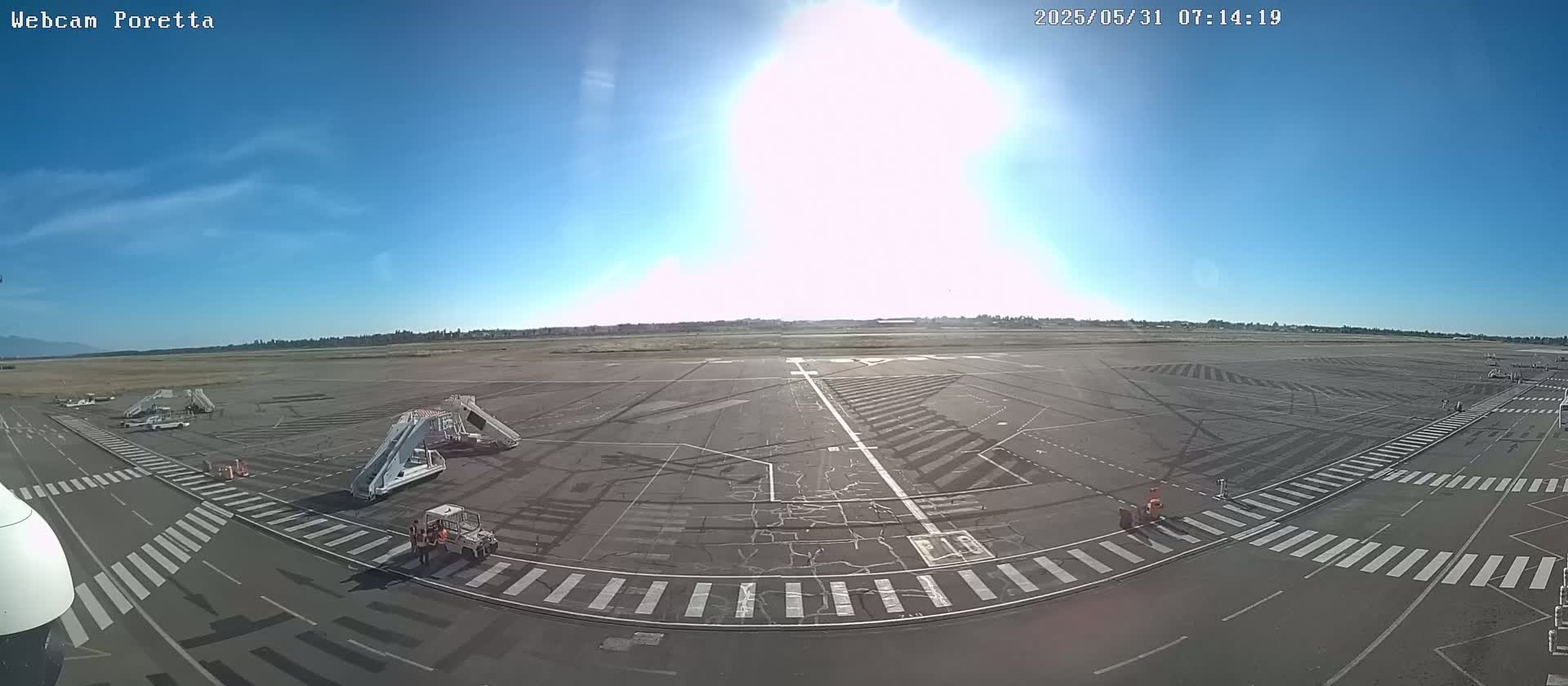 A sunny airport tarmac with several jet bridges and ground crew visible, under a mostly clear blue sky.