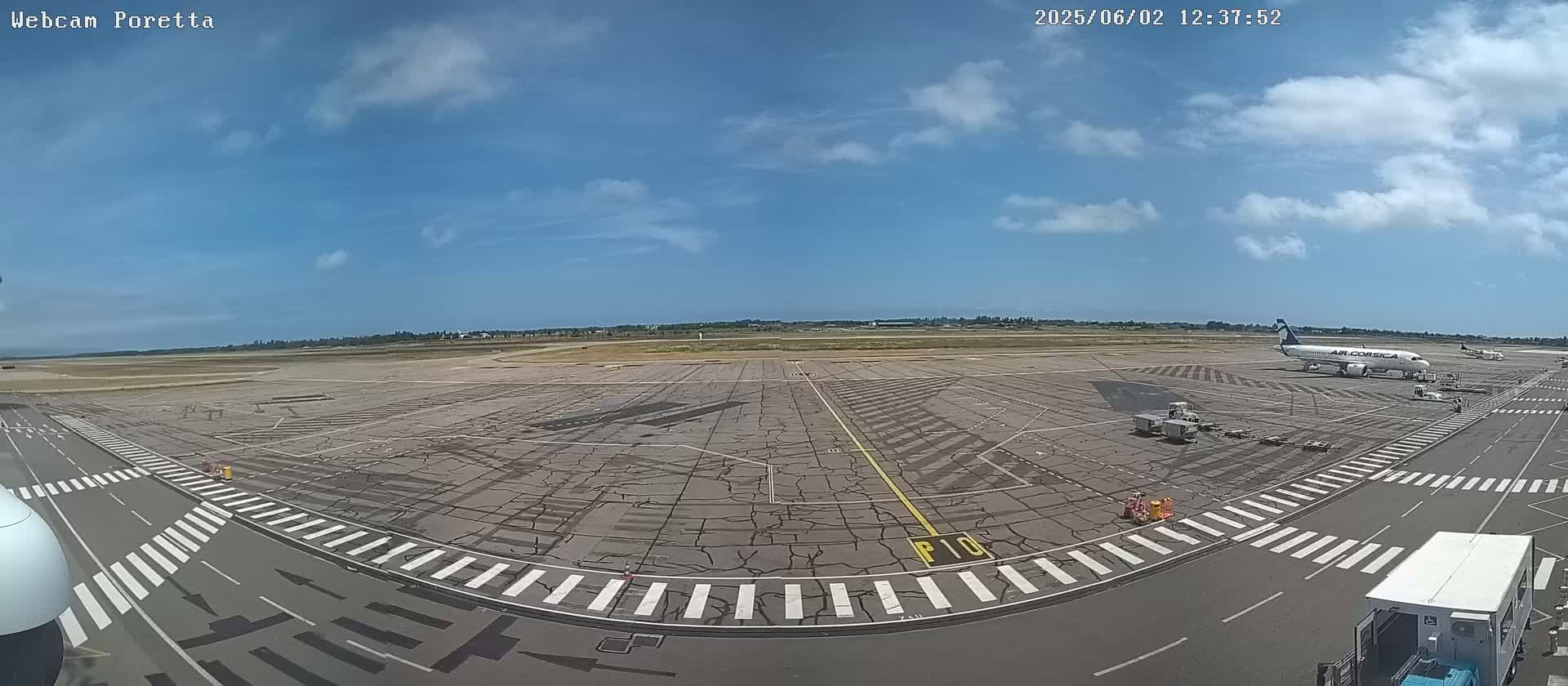A mostly sunny day at an airport tarmac shows an airplane parked near a service vehicle, with other vehicles and markings visible on the ground.