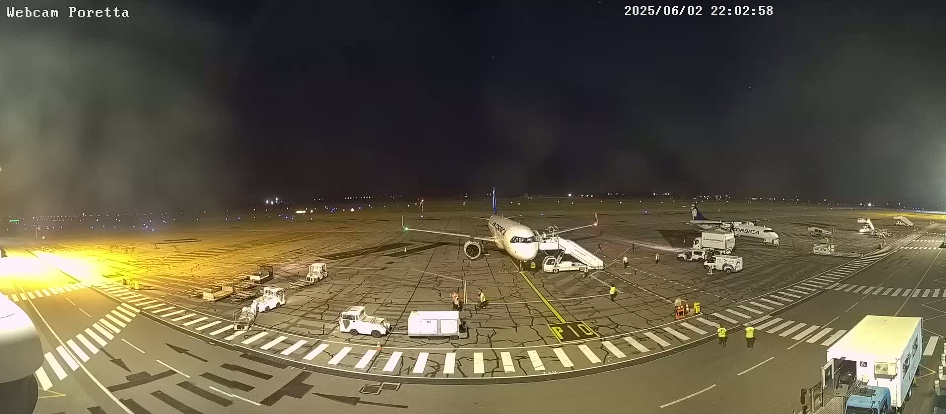 A passenger airplane is parked on an airport tarmac at night, with ground crews and service vehicles surrounding it under a hazy sky.