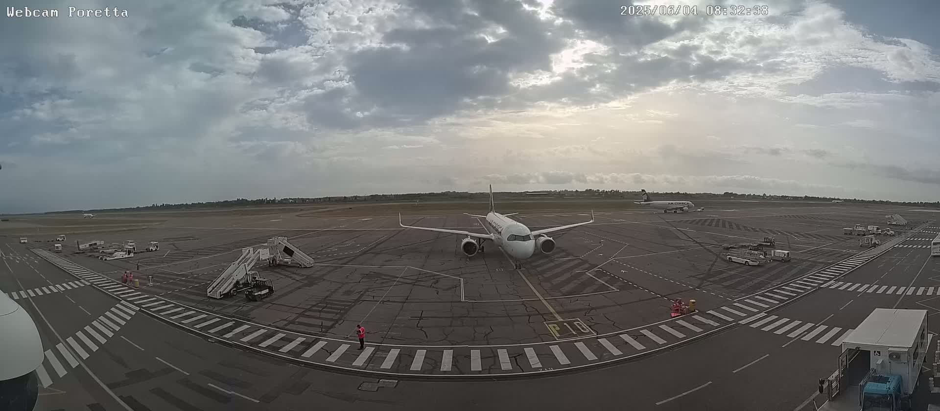 A mostly cloudy sky overlooks an airport tarmac where two airplanes are parked, one with a jet bridge attached, and various ground support vehicles are present.