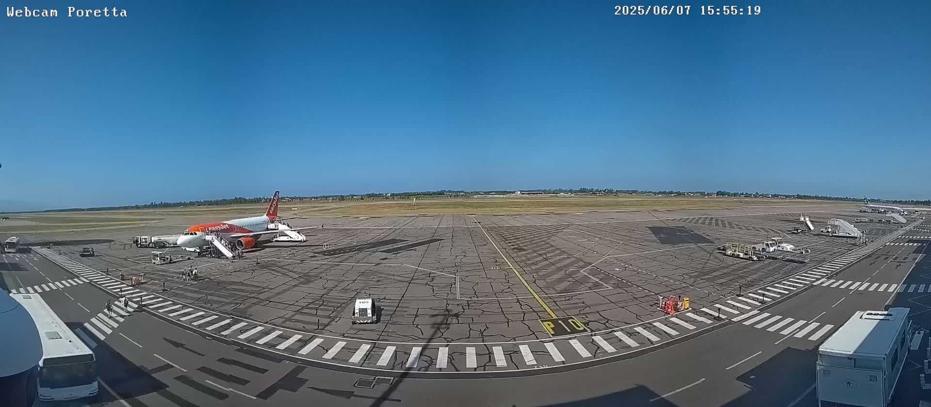 An airport tarmac on a sunny day shows an airplane at a gate with ground support vehicles and buses nearby.