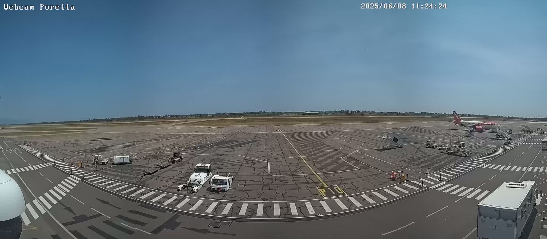 A high-angle, wide shot of an airport tarmac on a sunny day shows an airplane parked at a gate with several ground support vehicles present.