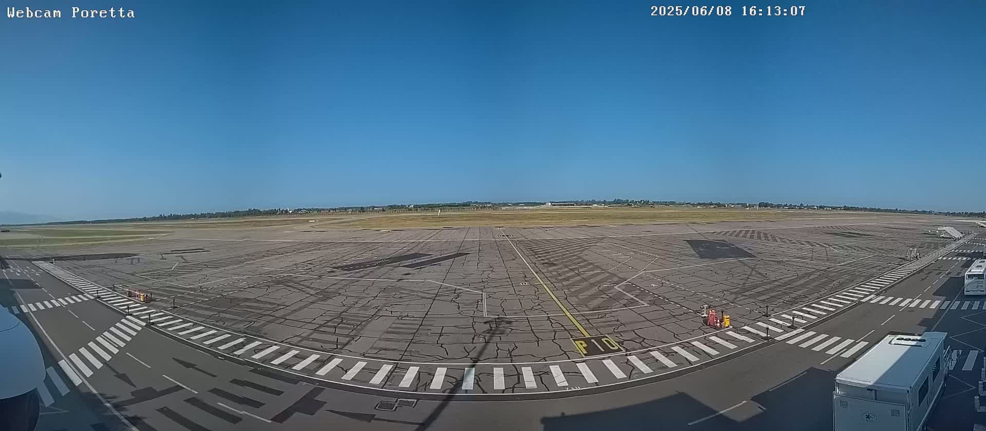 A wide shot of an airport tarmac on a clear, sunny day, showing various ground markings and some airport service vehicles.