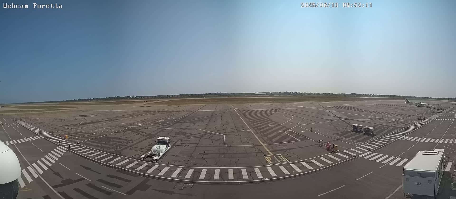 A wide, sunny view of an airport tarmac shows several airport service vehicles and a large white bus, with a single airplane visible in the distance.