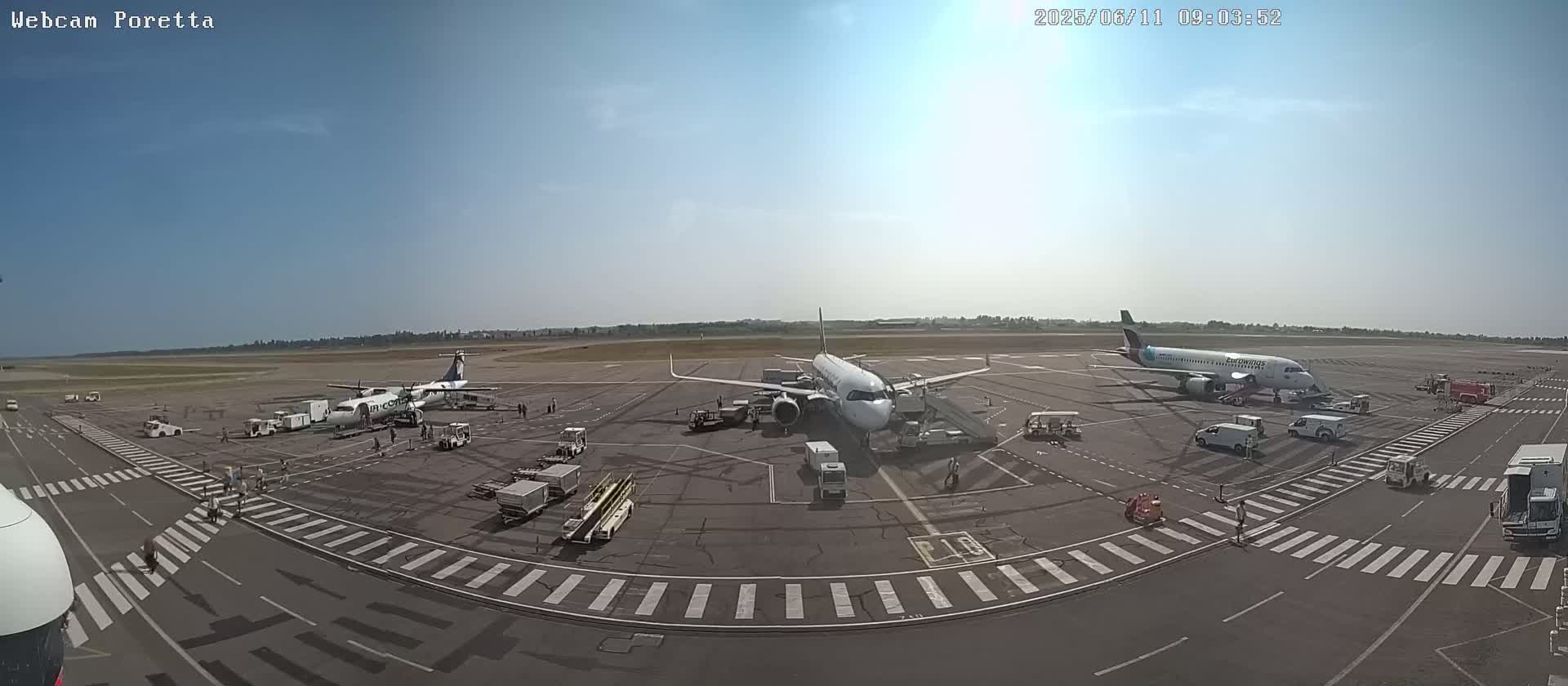 A sunny airport tarmac shows three airplanes parked near gates with various ground support equipment and personnel.