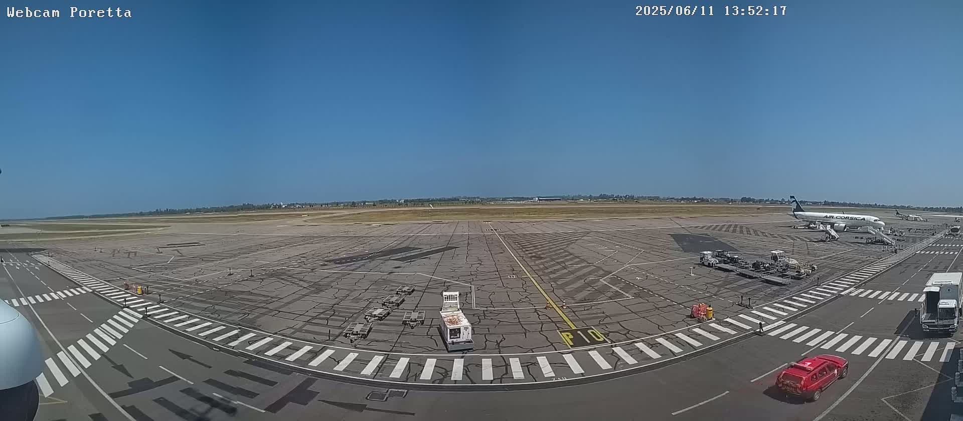 A wide, high-angle view of an airport tarmac on a sunny day shows an airplane at a gate with baggage carts and other ground support equipment surrounding it, along with several vehicles and crosswalks on the tarmac.