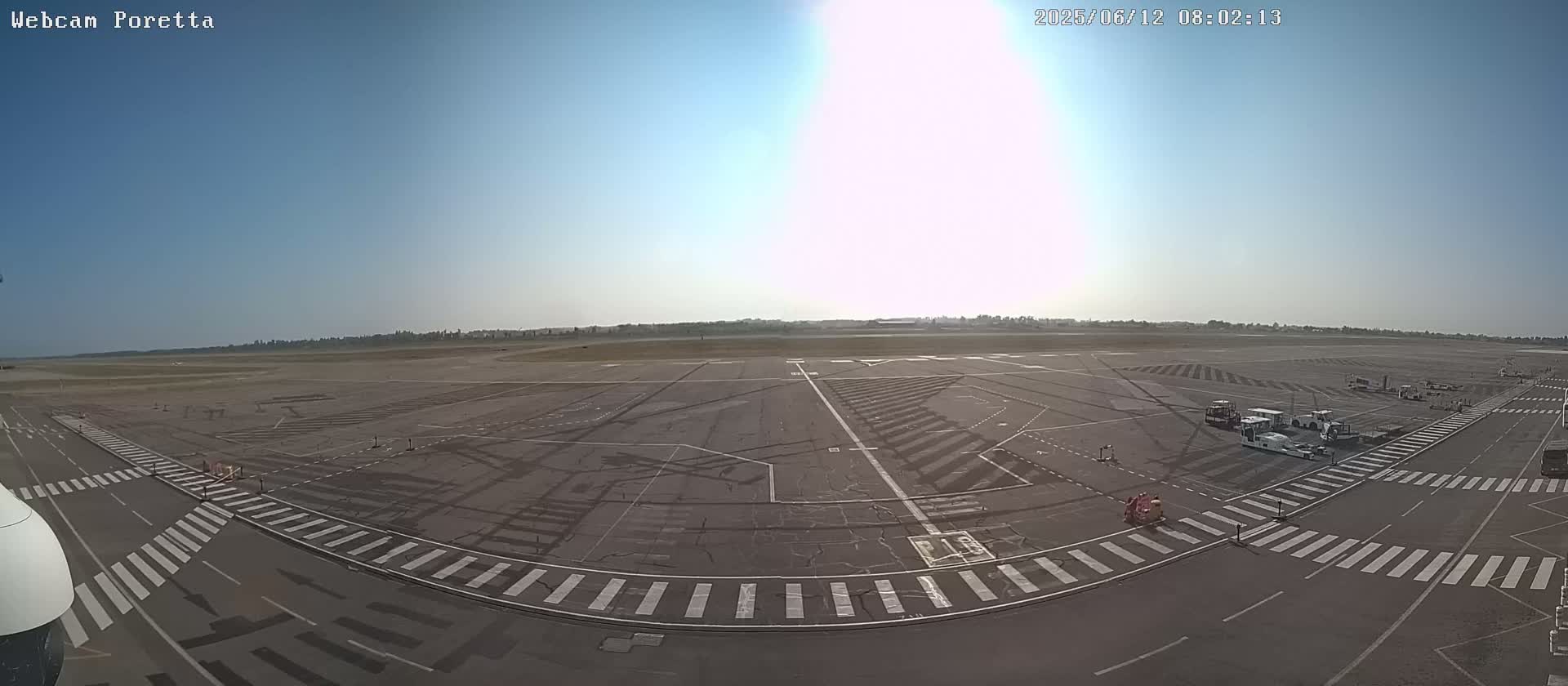 A mostly empty airport tarmac on a sunny day with several airport vehicles parked near a crosswalk.