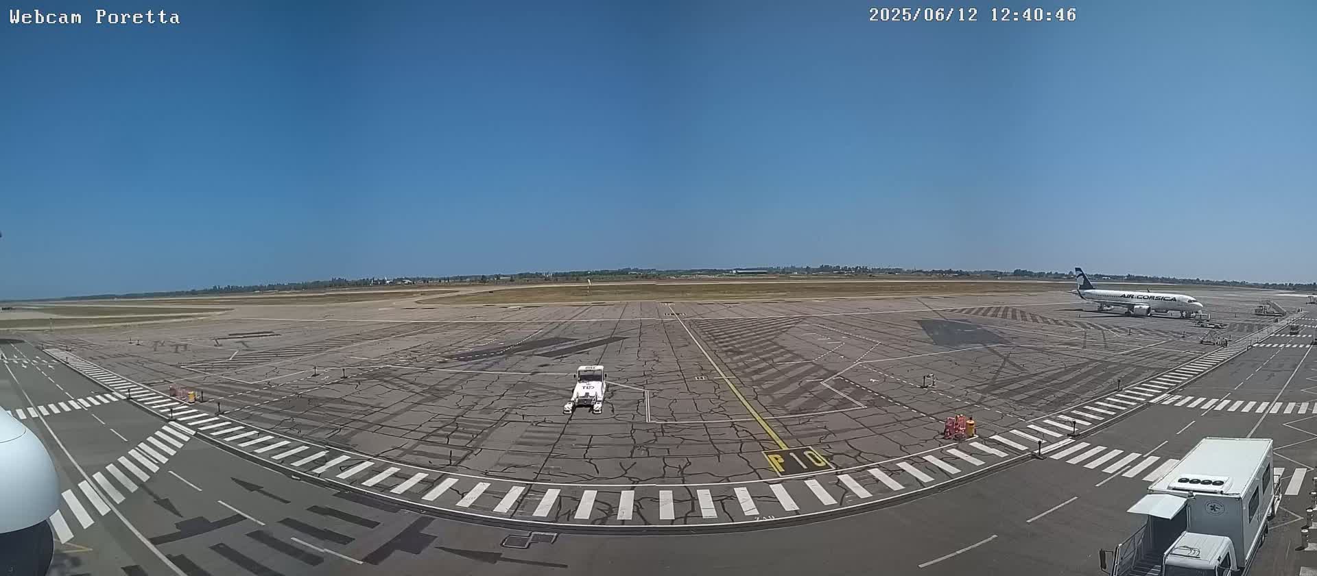 A wide shot of an airport tarmac on a sunny day shows a plane parked at the gate, airport service vehicles, and crosswalks on the pavement.