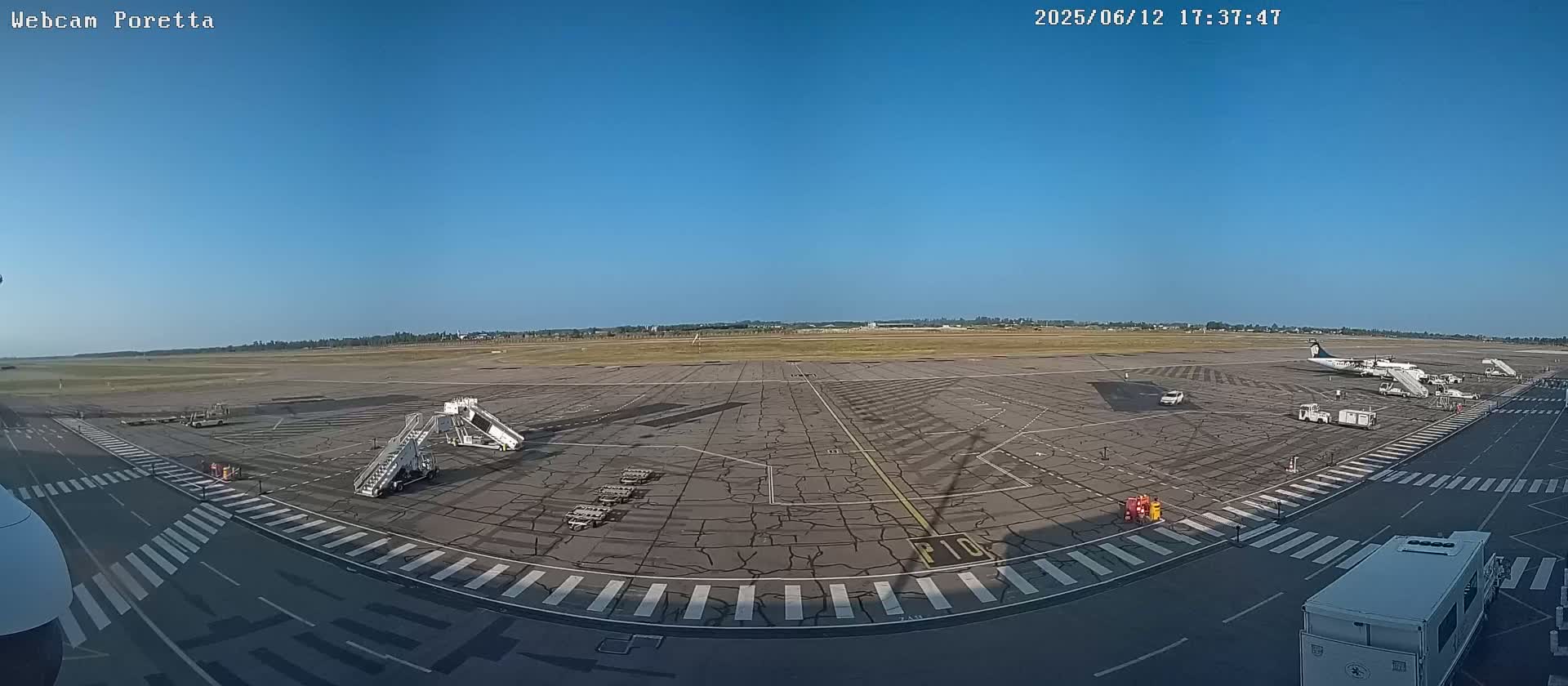 A wide shot of an airport tarmac on a sunny day shows several airplanes parked at gates, along with ground support equipment and jet bridges.
