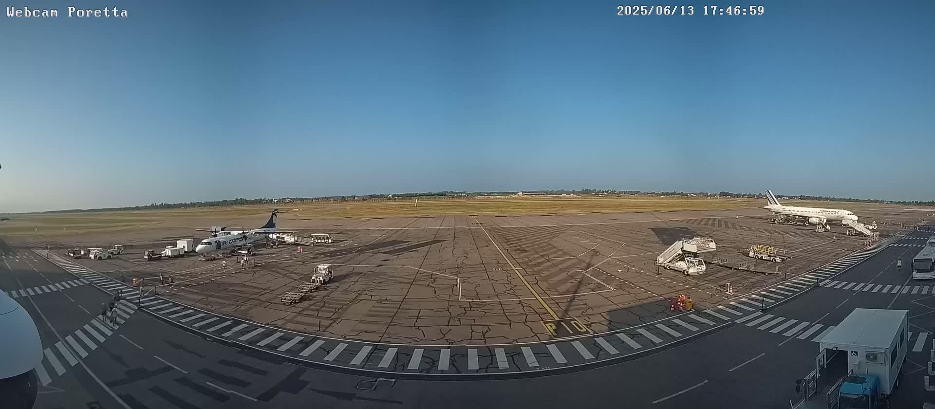 A mostly sunny airport tarmac shows two airplanes, several ground support vehicles, and crosswalks.