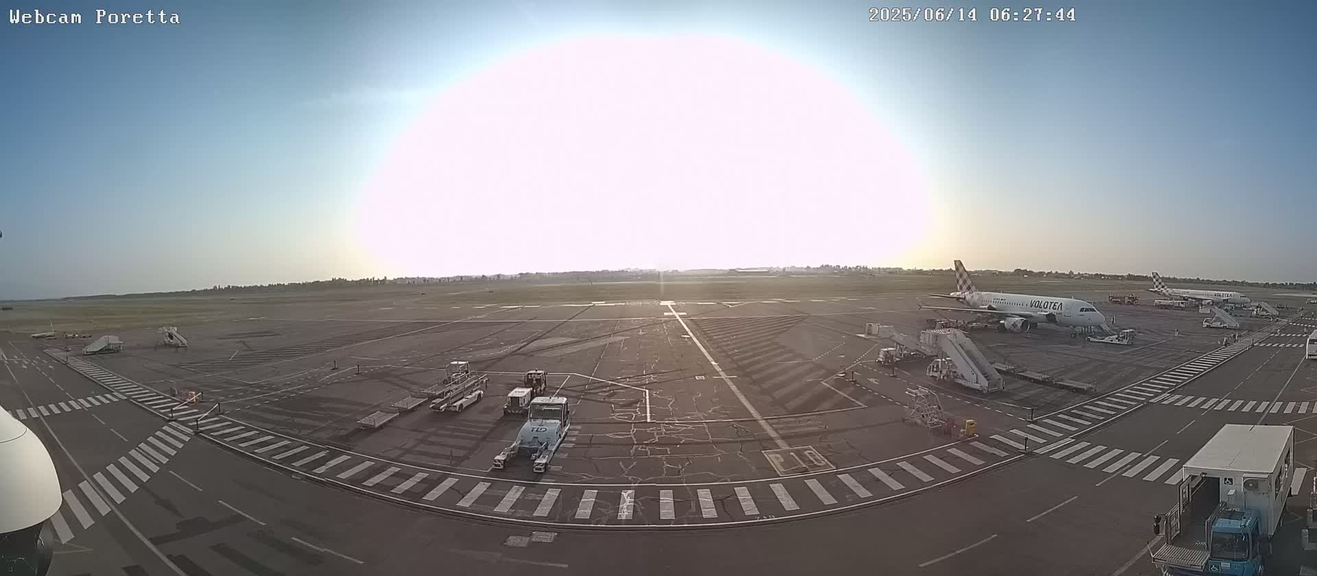 A mostly sunny airport tarmac shows two airplanes, several ground support vehicles, and crosswalks.