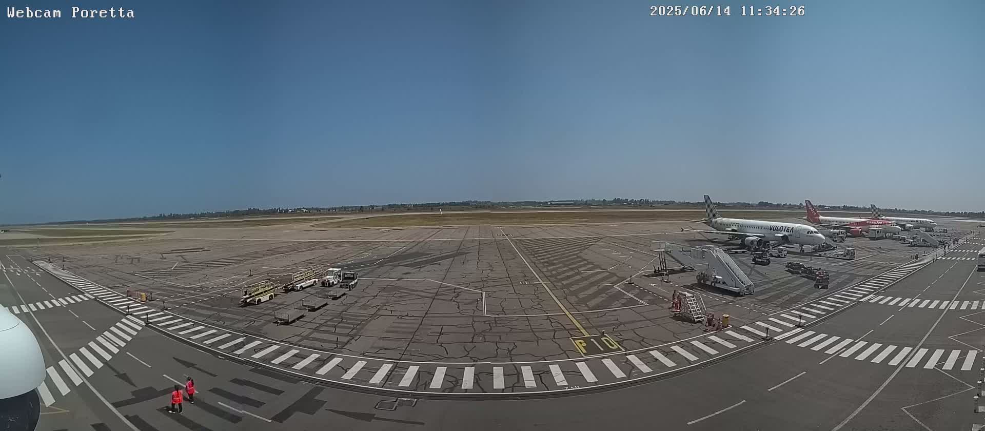 Several airplanes are parked on a tarmac under a clear blue sky, with ground support equipment and personnel visible.