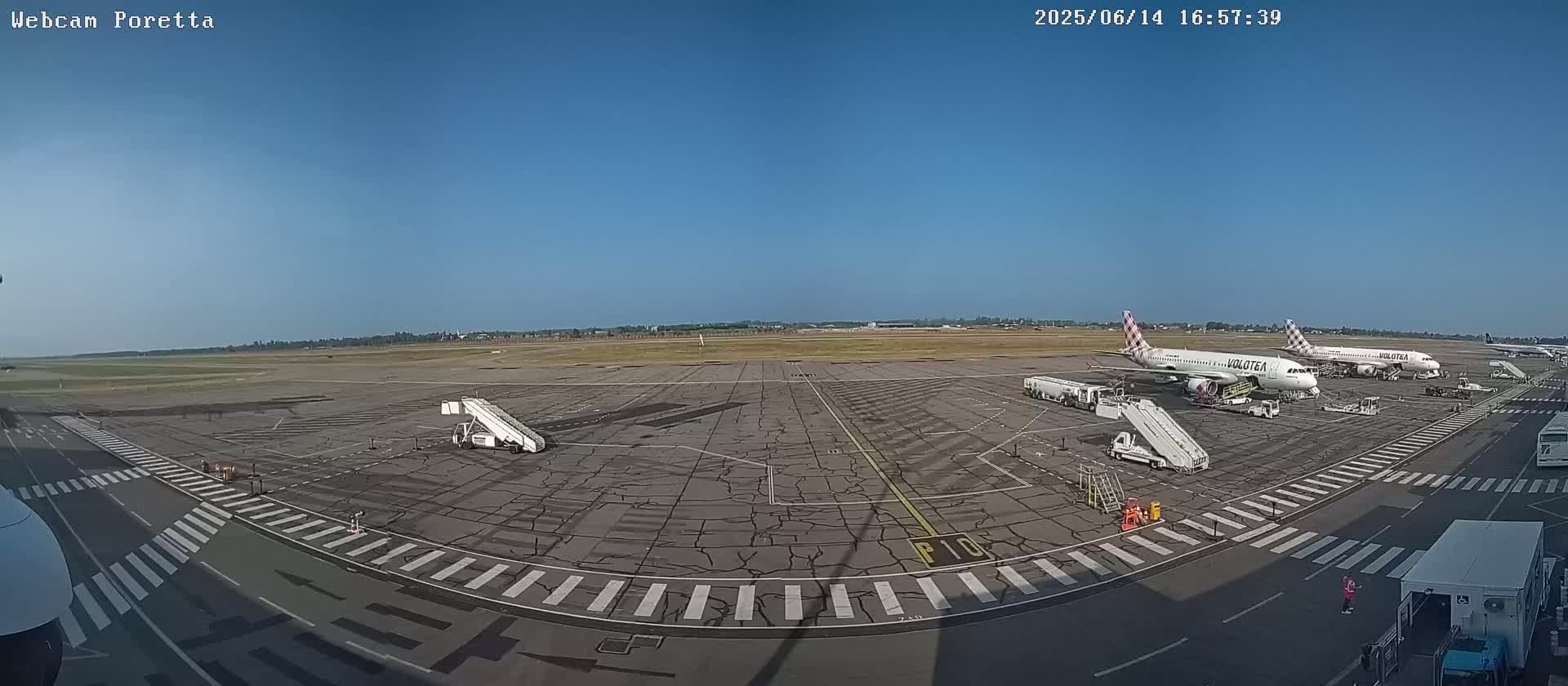 A wide outdoor shot on a clear sunny day shows several airplanes parked on a tarmac at an airport, with jet bridges and ground support equipment visible.
