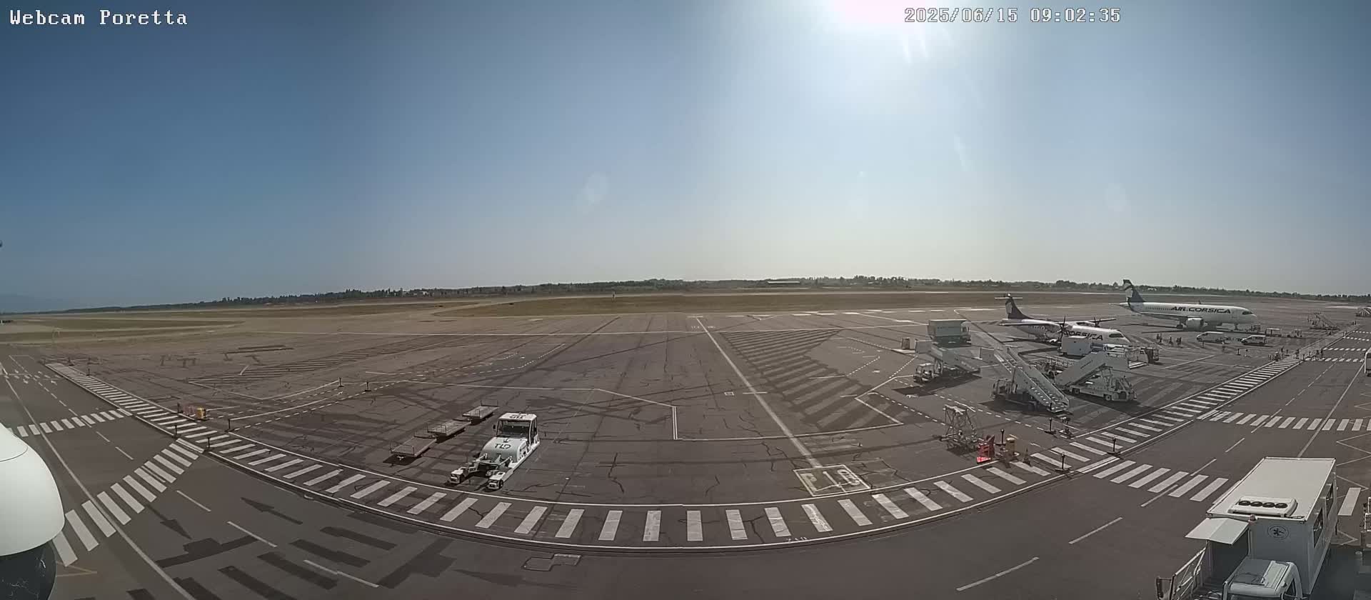 An airport tarmac on a sunny day with two airplanes parked at gates, baggage carts, and ground support equipment.