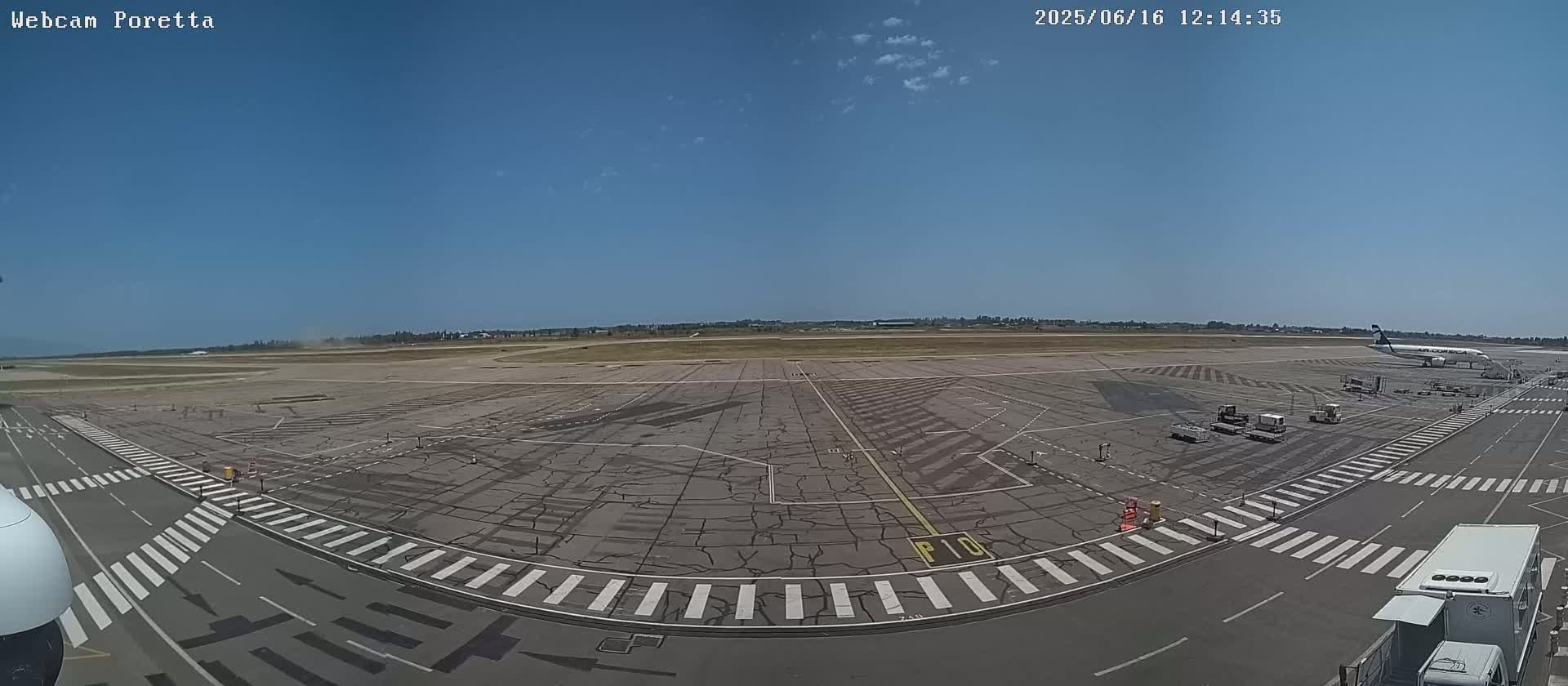 A mostly sunny airport tarmac with a single airplane parked at a gate and several ground service vehicles.