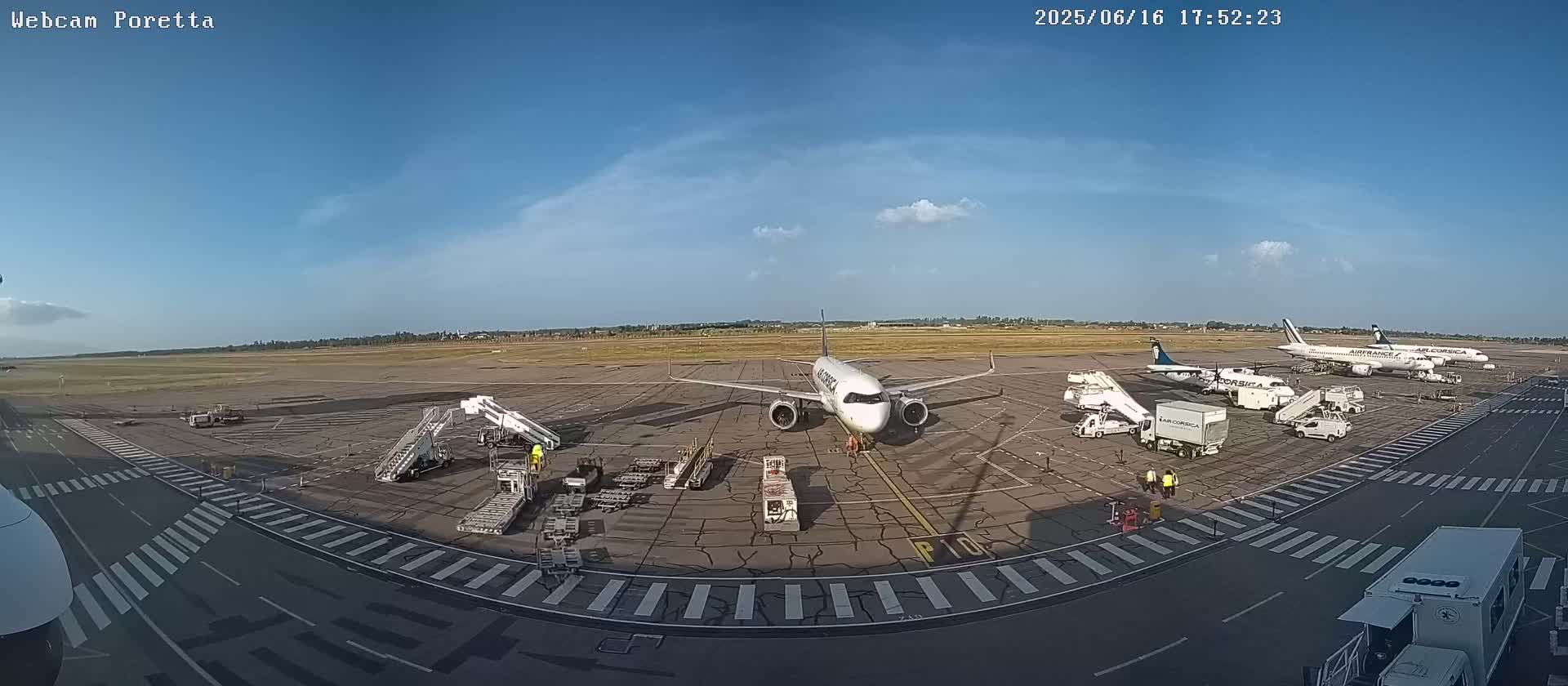 An airport tarmac under a mostly clear blue sky shows several airplanes at gates with ground support equipment and personnel.