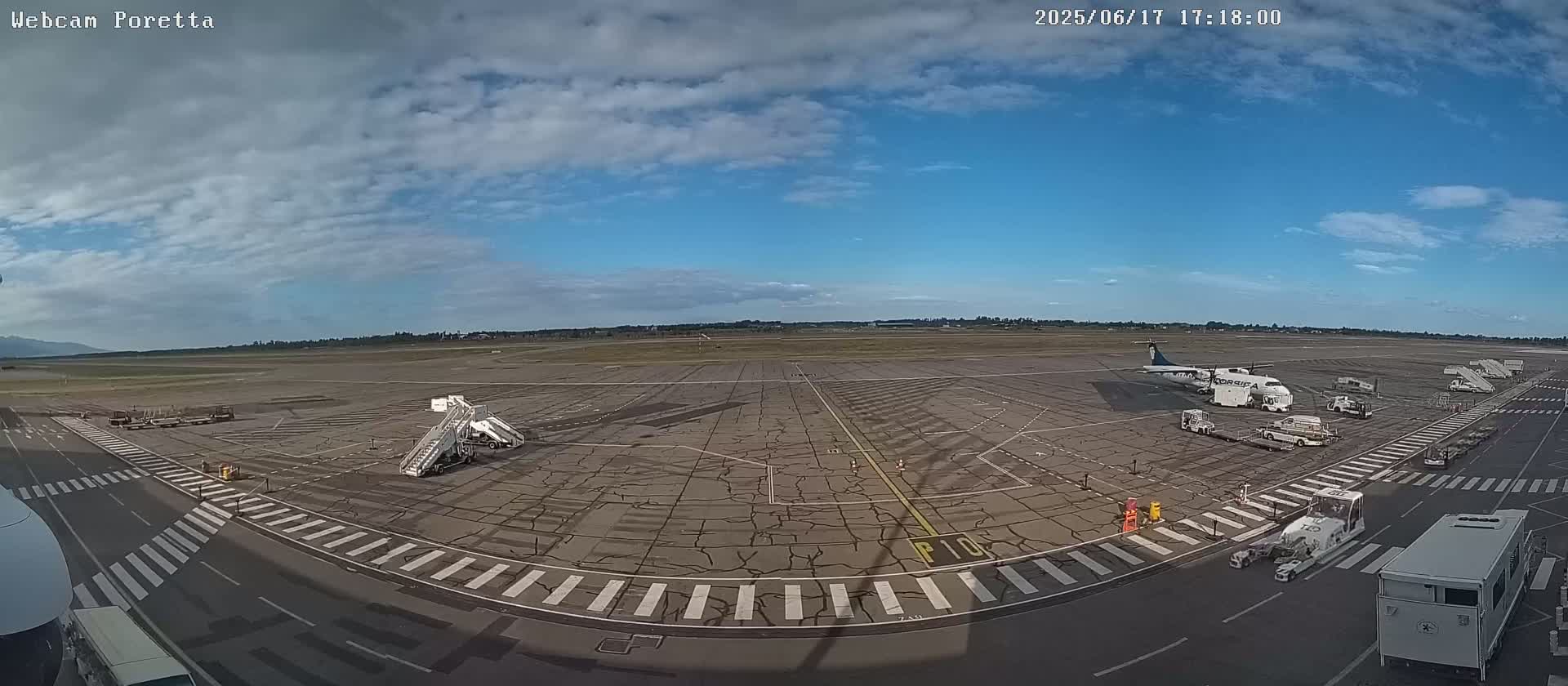 A mostly sunny airport tarmac with several airplanes, jet bridges, and ground support vehicles visible under a partly cloudy sky.