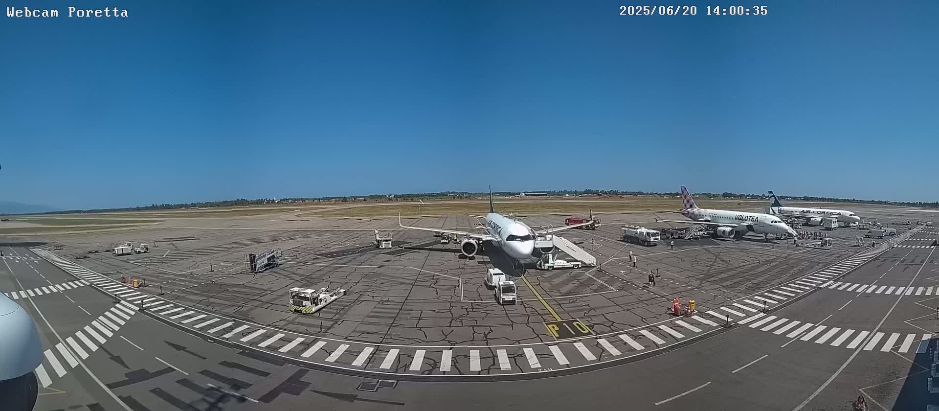 A wide shot of an airport tarmac on a sunny day shows three airplanes parked at gates with ground crews and equipment present.