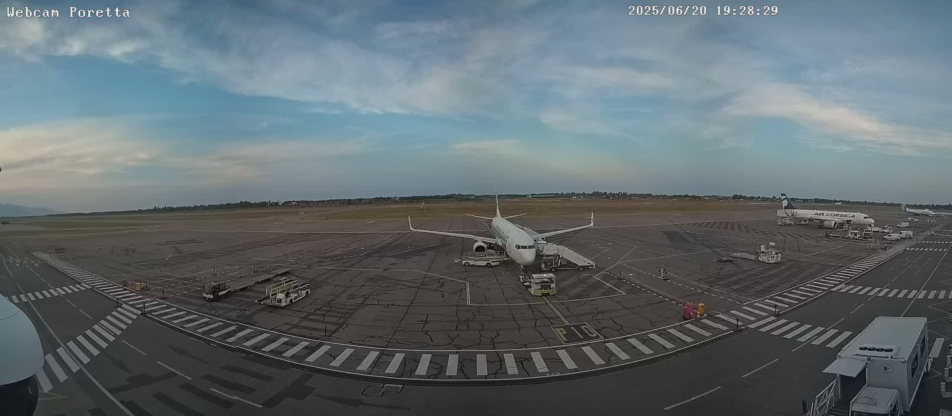A wide view of an airport tarmac under a partly cloudy sky shows several airplanes and ground support vehicles.