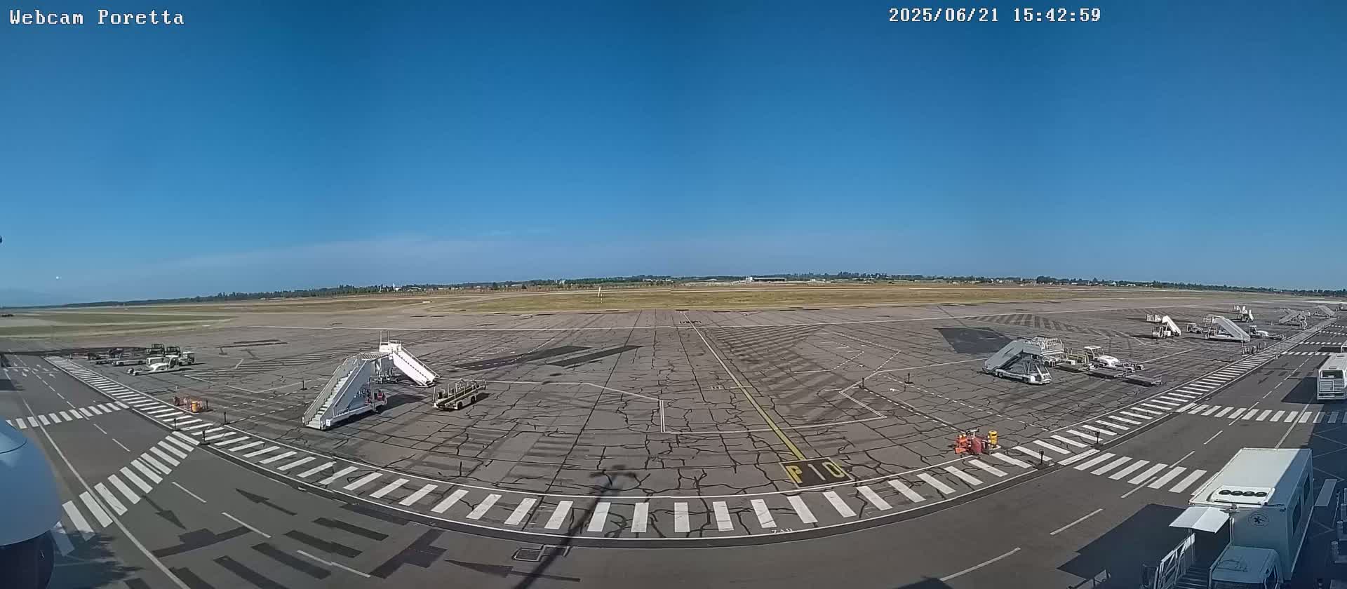 A wide shot of an airport tarmac on a sunny day shows several jet bridges and ground support equipment parked near the terminal building.