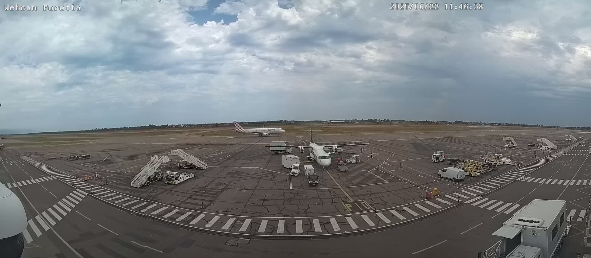 An airport tarmac under a cloudy sky shows several airplanes, support vehicles, and passenger boarding bridges.
