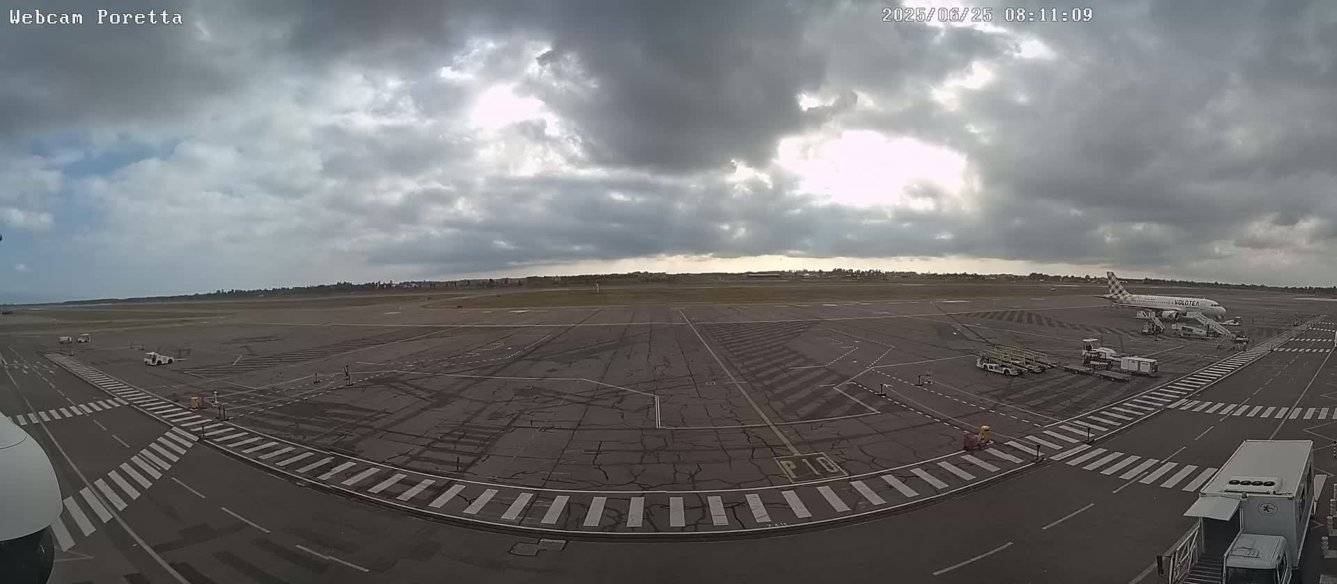 A mostly cloudy sky hangs over an airport tarmac where a passenger plane is parked at a gate, surrounded by ground support vehicles.