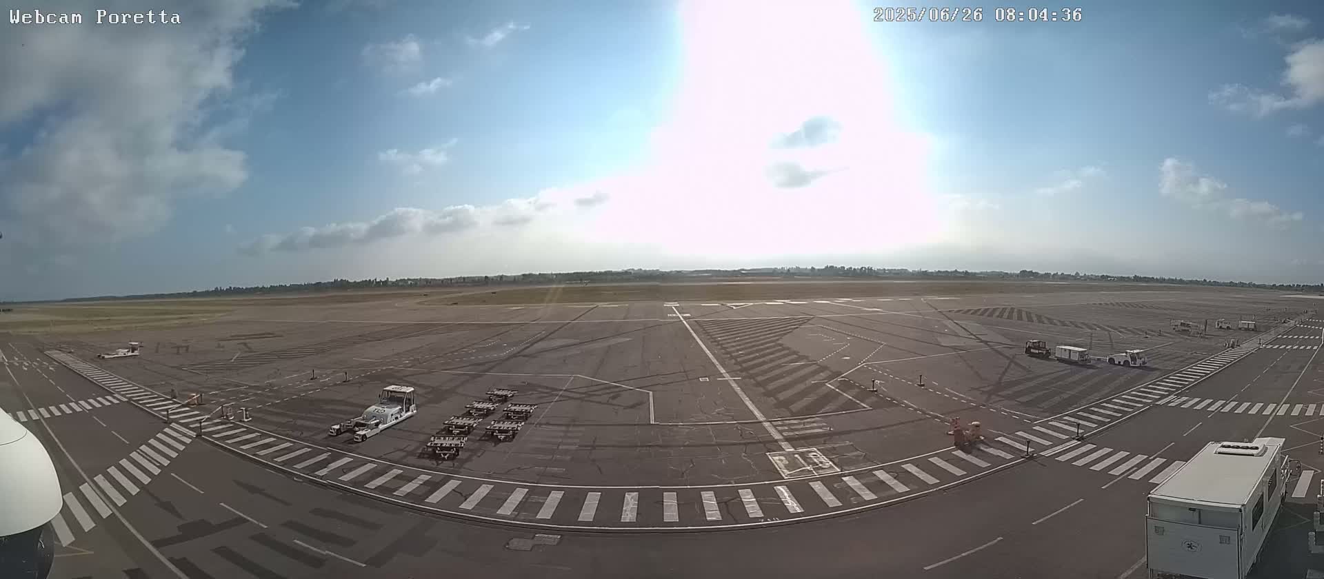 A mostly sunny day at an airport tarmac shows several ground support vehicles and baggage carts parked on the runway.