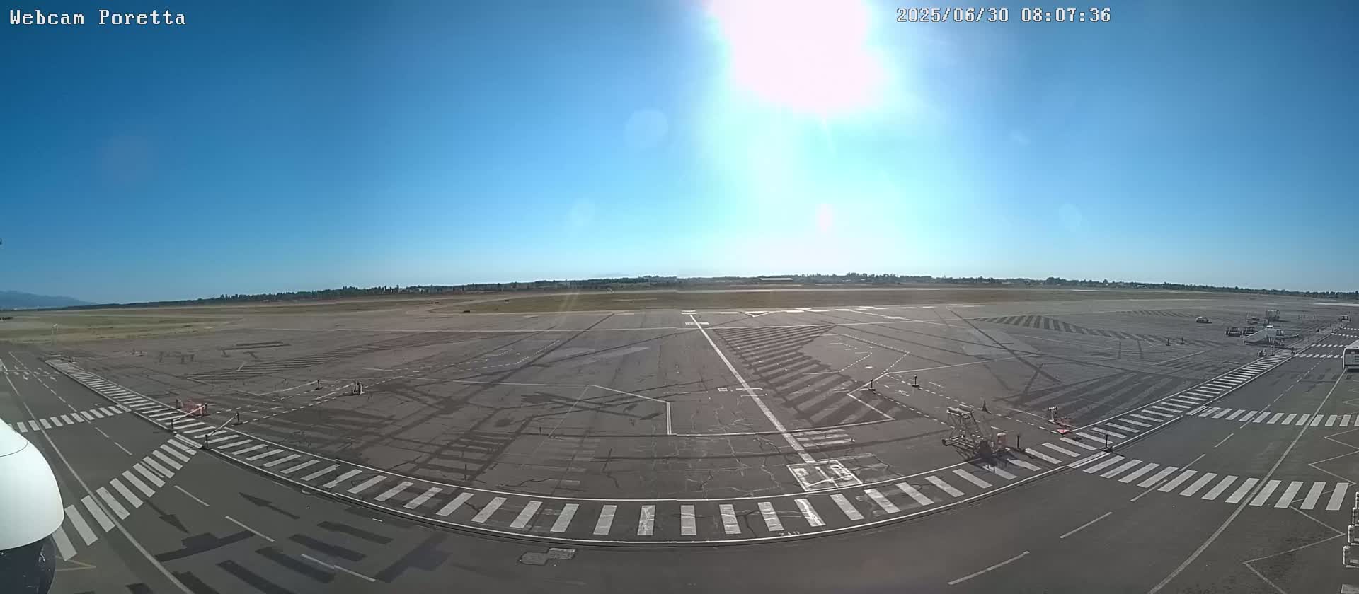 A sunny day at an airport tarmac shows numerous markings on the ground, with several airport service vehicles visible in the distance.