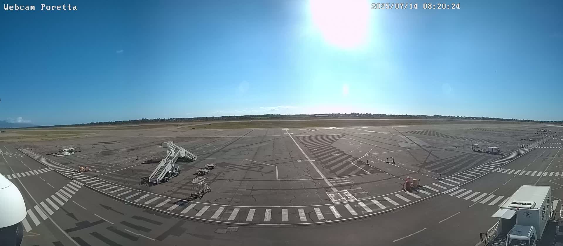 A sunny airport tarmac is shown with jet bridges, service vehicles, and crosswalks.