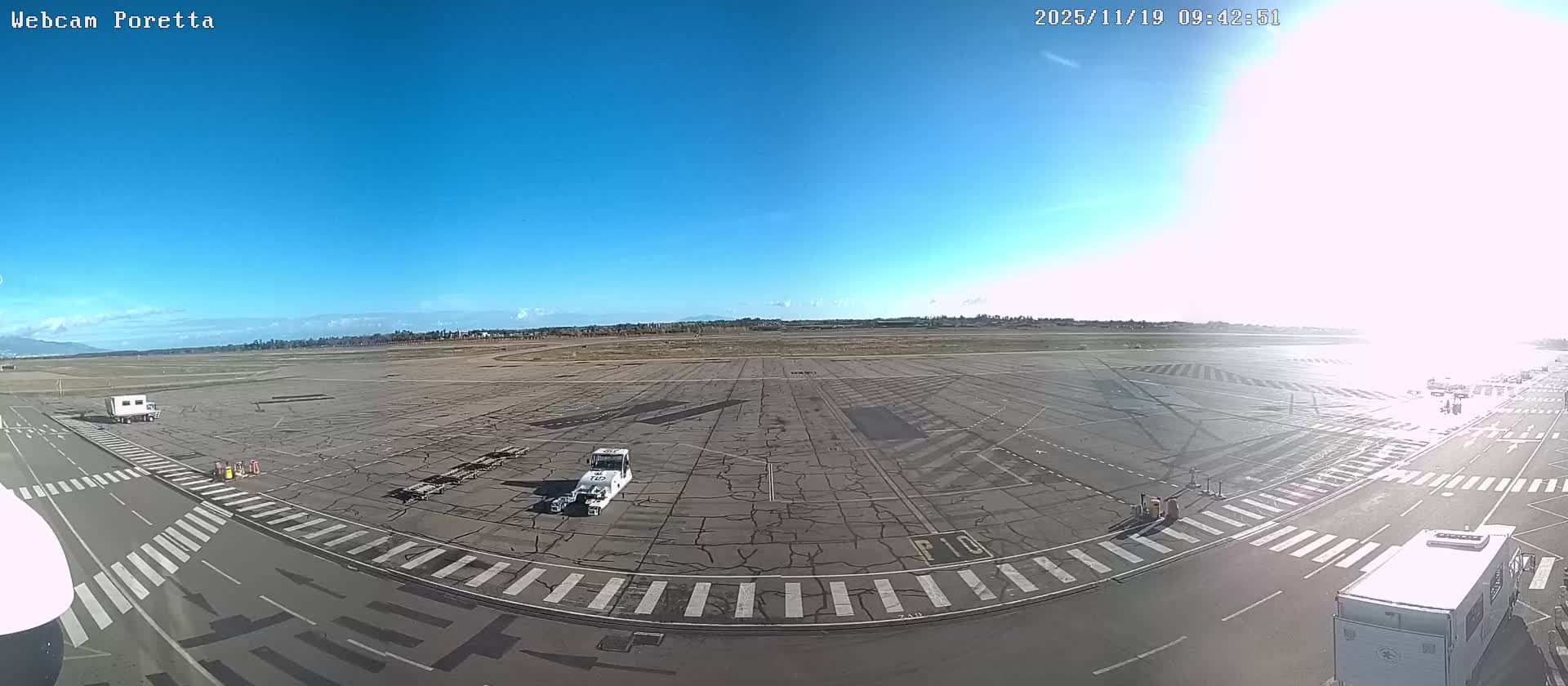 An airport tarmac with ground support vehicles and markings stretches under a bright, clear blue sky on a sunny day, with a strong sun glare overexposing the right side of the scene.