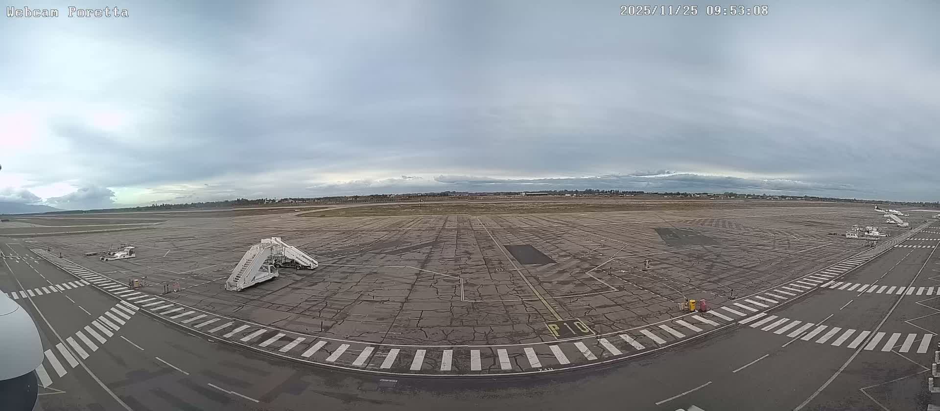 The image depicts a wide airport tarmac with ground support equipment and distant parked aircraft under a cloudy, overcast sky, with mountains visible on the horizon.