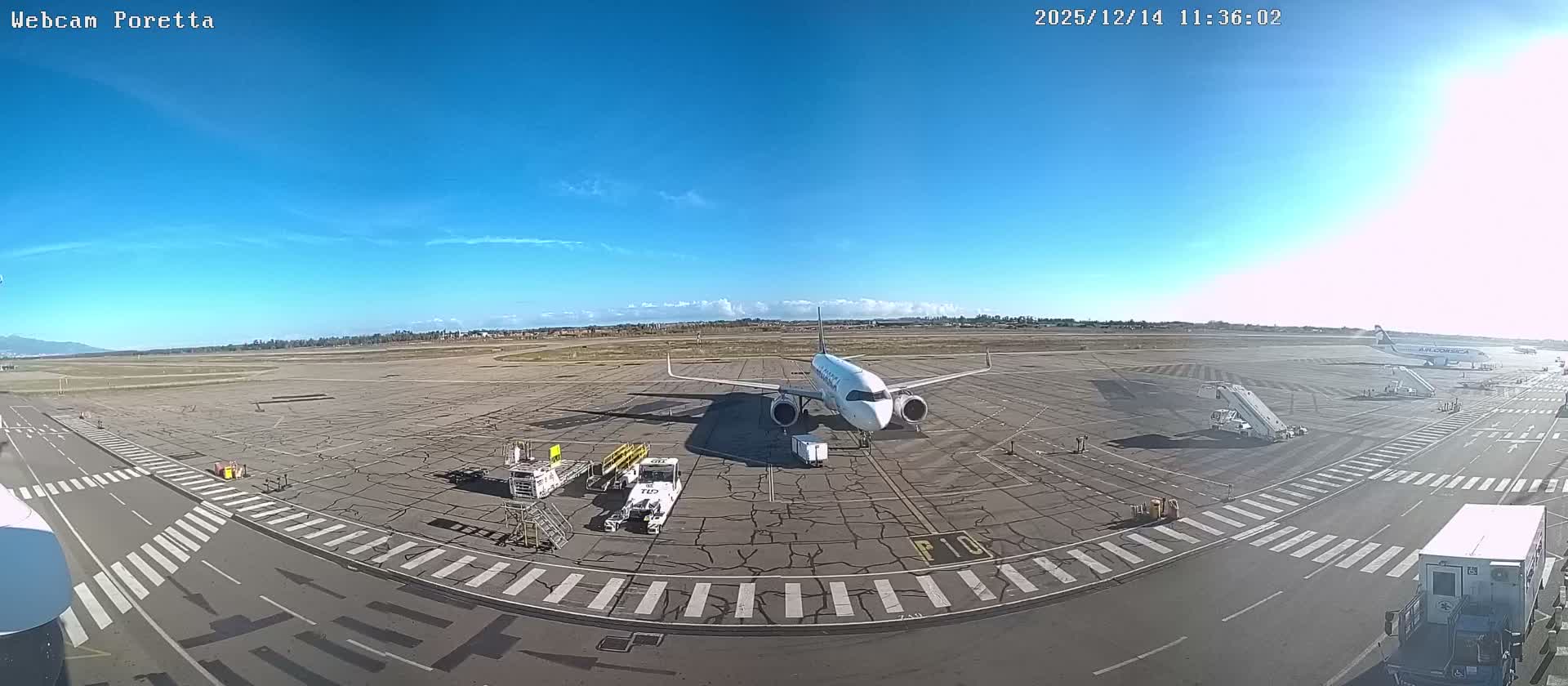 The image depicts a wide airport tarmac with ground support equipment and distant parked aircraft under a cloudy, overcast sky, with mountains visible on the horizon.