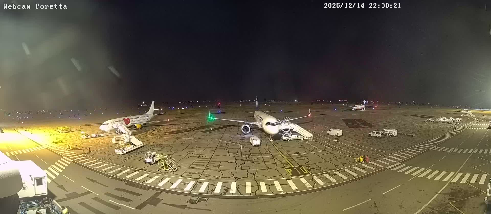The image depicts a wide airport tarmac with ground support equipment and distant parked aircraft under a cloudy, overcast sky, with mountains visible on the horizon.