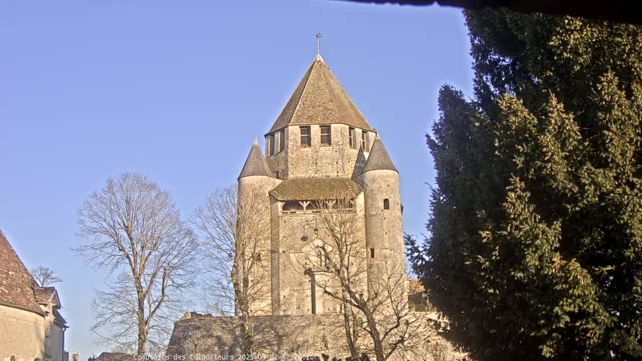 A tall stone tower with a conical roof stands among bare trees under a clear blue sky.