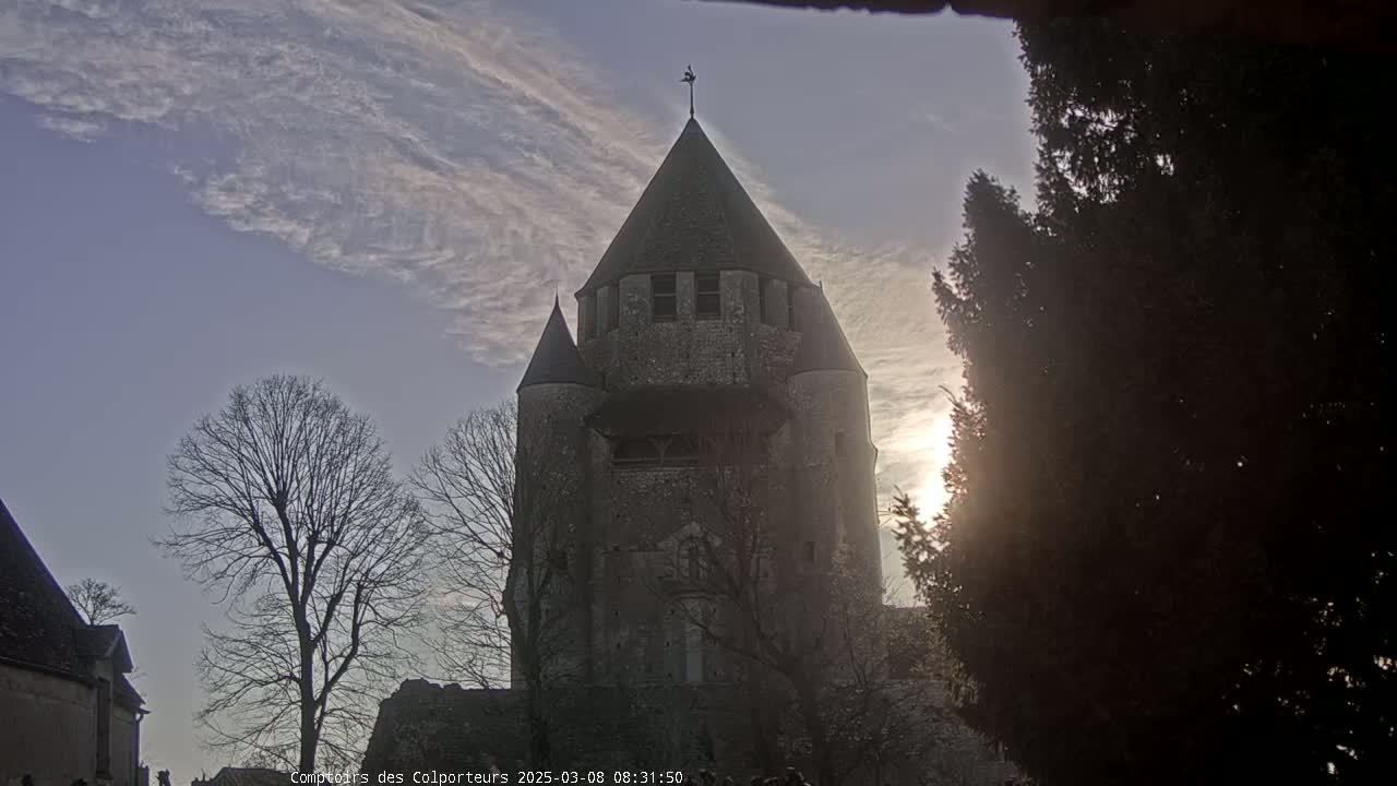 A tall, stone tower with conical roofs is seen under a partly cloudy sky, backlit by the low sun.