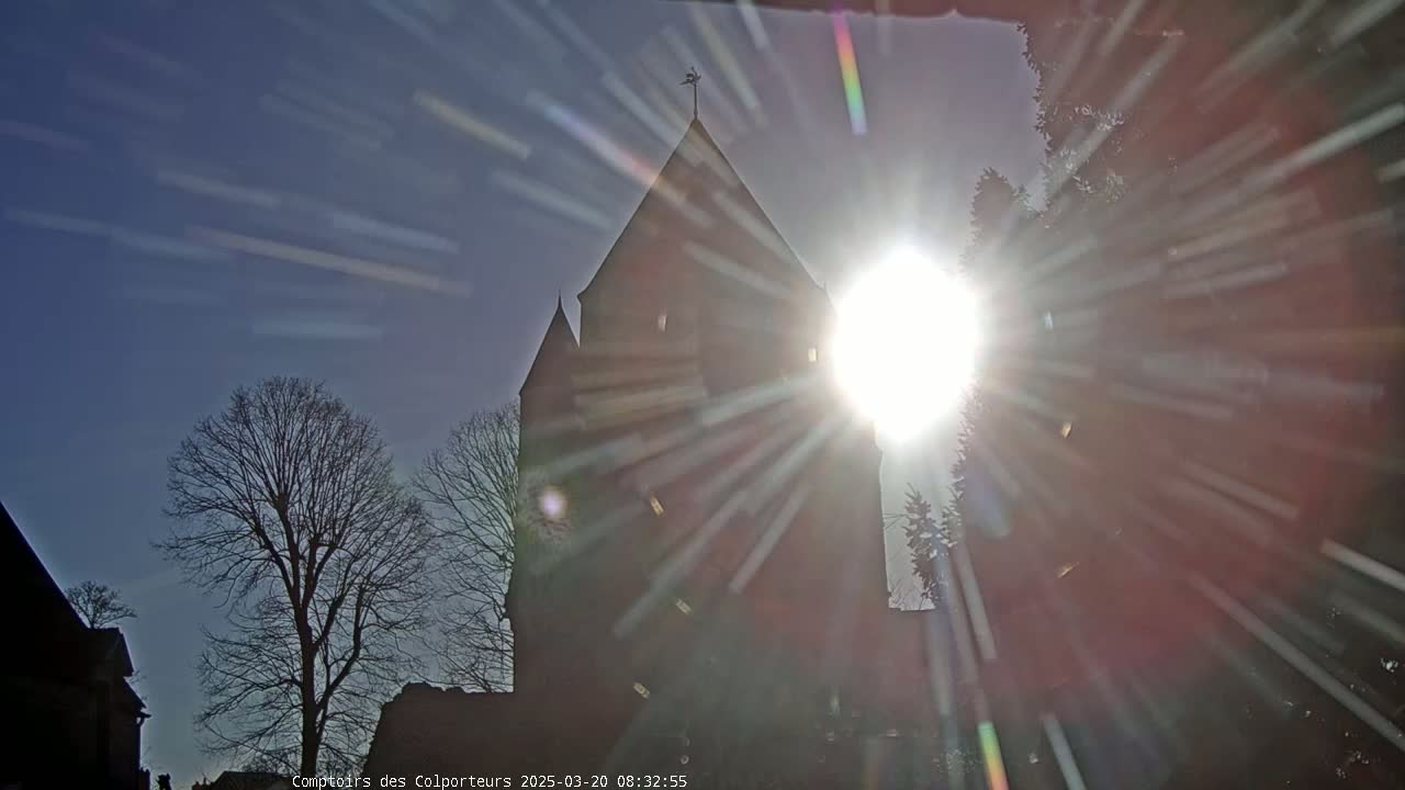 A tall, dark church steeple is silhouetted against a bright sun with starburst rays, in a clear sky.