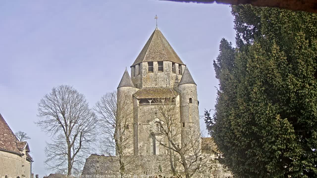 A large stone tower, with a conical roof and smaller flanking towers, stands amidst bare deciduous trees and an evergreen tree under a pale blue sky.